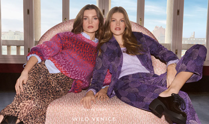 2 Frauen sitzen in einem hellrosa Sessel. Eine Frau trägt einen Rock mit Leopardenprint, ein hellblaues Hemd und einen rosa Pullover. Die andere Frau trägt einen violetten Anzug mit Muster und schwarze Schuhe